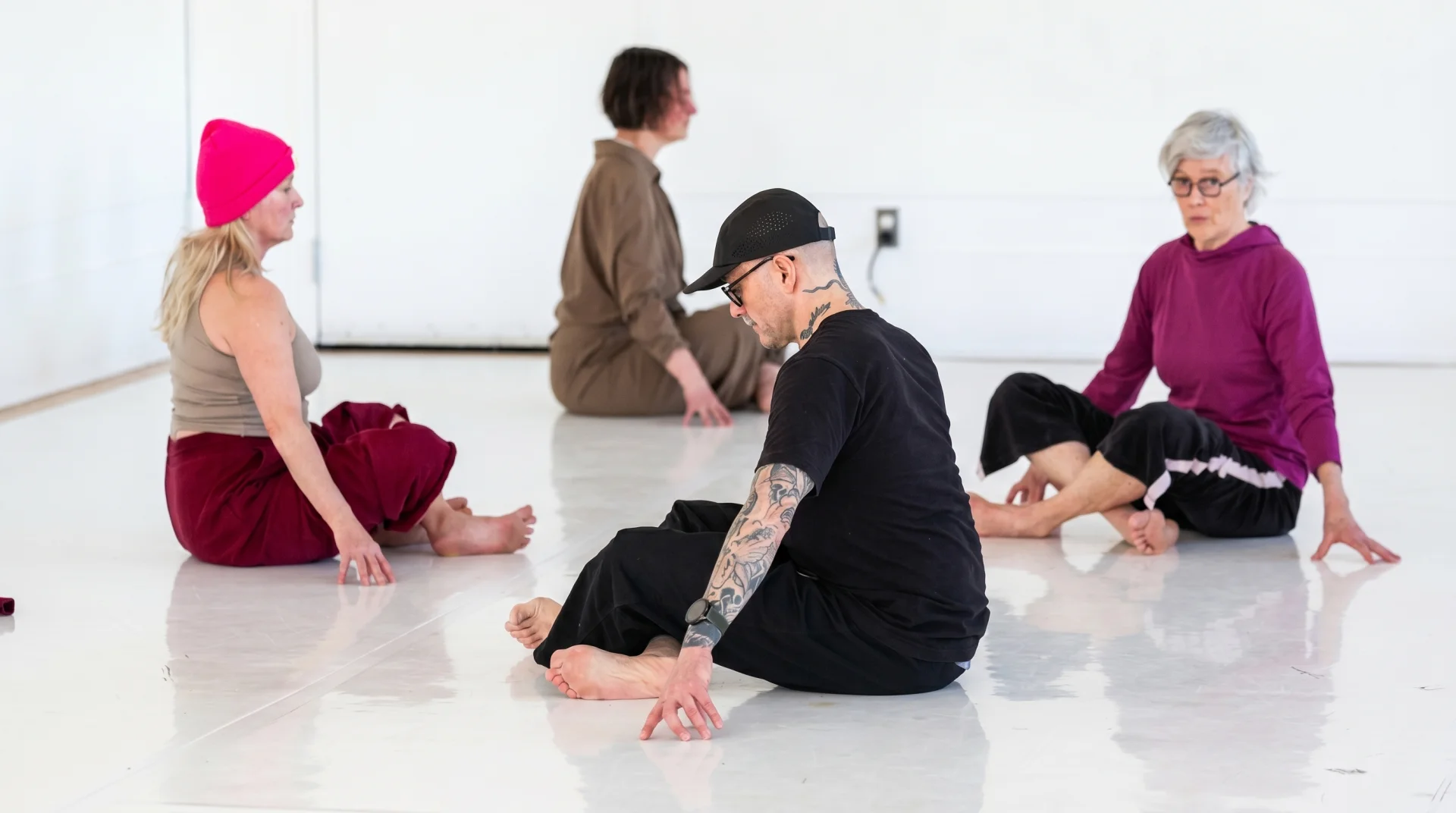 Four people seated on a white studio floor during a Baseworks practice session. A person at center wearing a black cap sits cross-legged with bowed head and hands resting on the floor, attention turned inward. Three others — in a pink hat, olive jumpsuit, and purple sweater — sit in similar settled postures around the space.