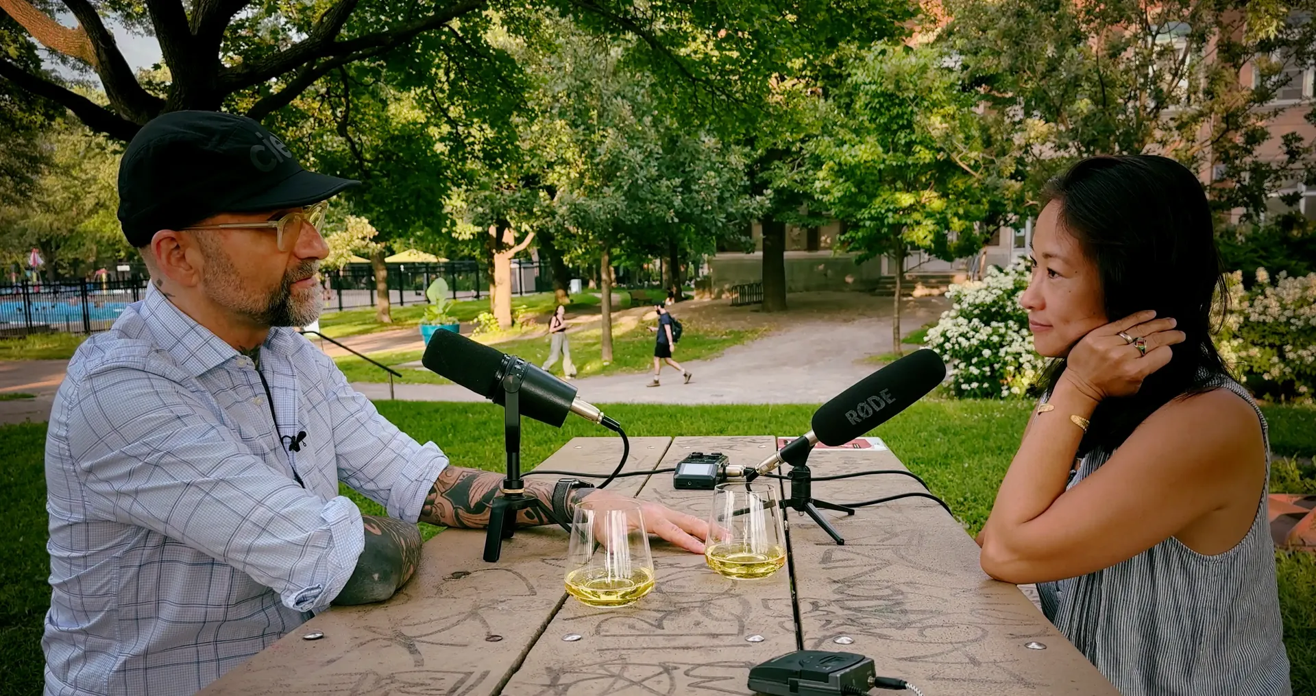 A man and a woman sitting in a park talking in a podcast.