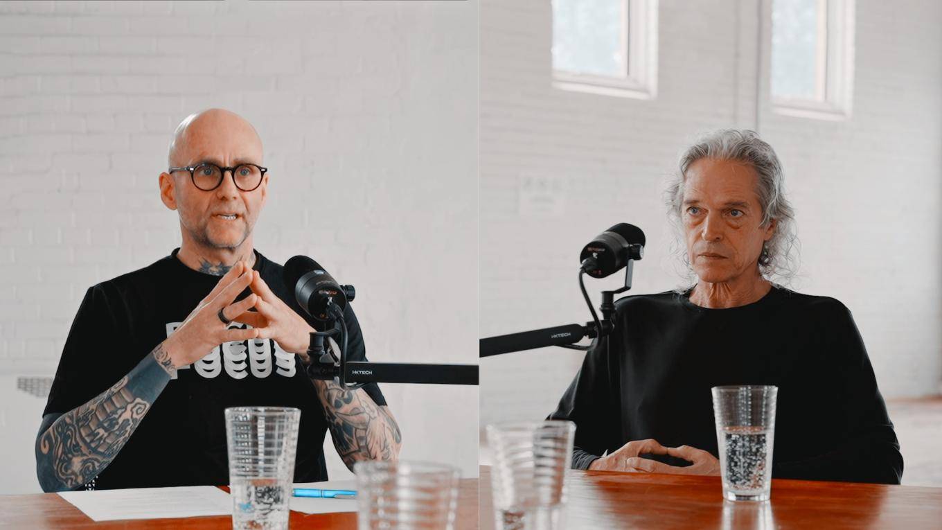 Two men speak into microphones at a table.